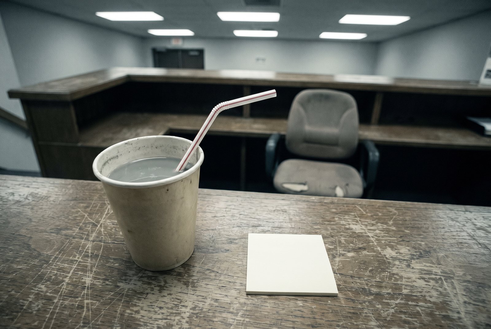 A paper cup of cloudy water with a bent straw on a scratched reception counter beside a blank note.